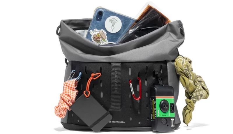 A gray and black camera bag filled with various items, including a tablet with stickers, a notebook, a bandana, a disposable camera, portable hard drive, and carabiners with keychains attached outside the bag.