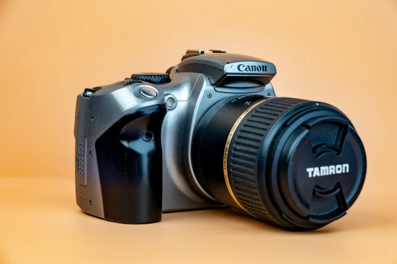 A Canon DSLR camera with a Tamron lens attached, placed on a smooth, light orange background. The camera is angled slightly to the left, prominently displaying the Canon and Tamron brand names.