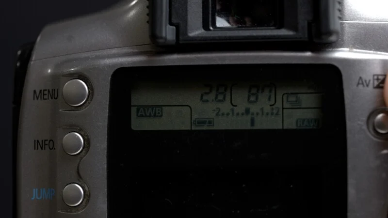 Close-up of a digital camera's control panel and LCD screen displaying camera settings. The screen shows “2.8 (87),” along with icons for AWB, battery level, and RAW format. Menu, info, and jump buttons are visible on the left.