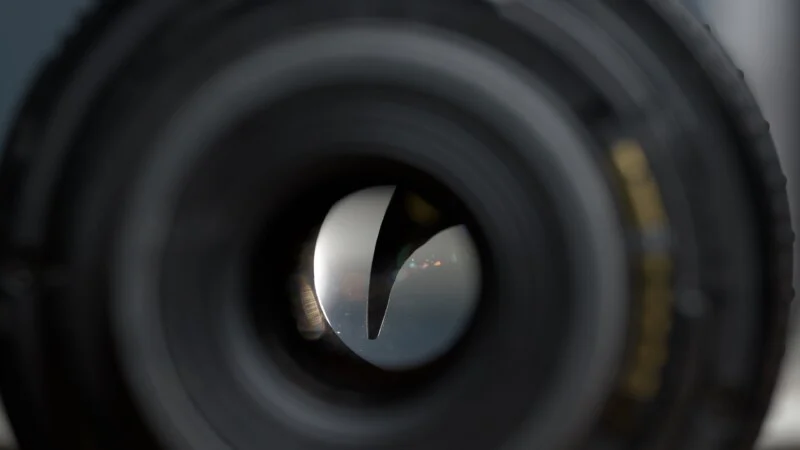 Close-up view of a camera lens aperture, showing the curved blades partially closed with reflections of light visible on the inner glass elements.