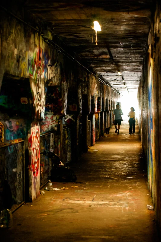 Two people walk toward the light at the end of a long, narrow tunnel covered in colorful graffiti, with dim overhead lights illuminating the passage.
