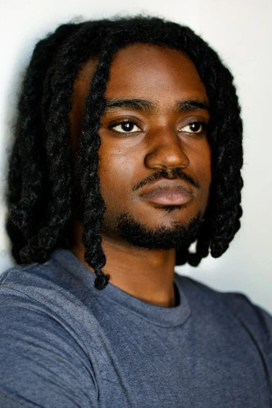 A person with medium-length twisted hair and a short beard is wearing a gray shirt, standing against a light background, looking slightly to the side with a serious expression.