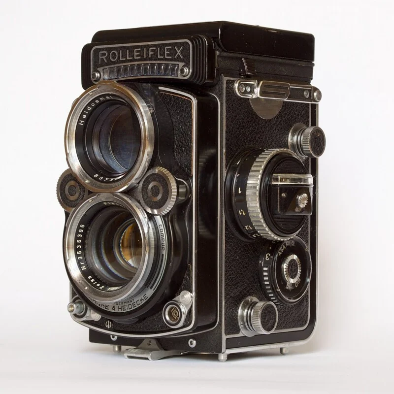 A vintage Rolleiflex twin-lens reflex camera with two large lenses, various dials, and a textured black body, displayed against a plain white background.