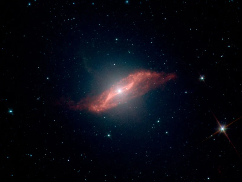 A bright spiral galaxy with a glowing, reddish-orange dust ring at its center, surrounded by numerous tiny white stars scattered across a dark space background.