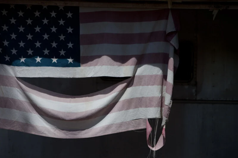 A faded and tattered American flag hangs, the bottom edge frayed and curling, with shadows cast across it. The background appears dark and metallic, suggesting an industrial or weathered setting.