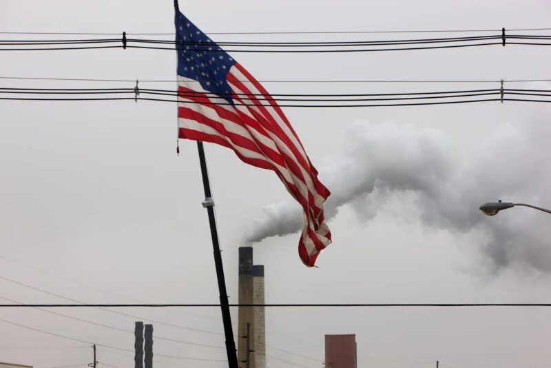 A tattered American flag hangs on a pole in the foreground, with industrial smokestacks in the background emitting plumes of smoke into a gray sky, intersected by several power lines.