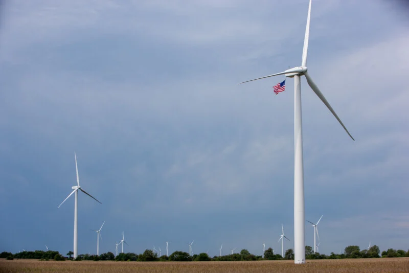 Wind turbines stand in a field under a blue sky, with an American flag attached to the blade of the nearest turbine. Trees and more turbines are visible in the background.