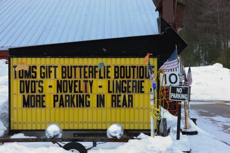 A yellow sign reads “Toms Gift Butterflie Boutique DVD’s - Novelty - Lingerie More Parking in Rear.” Nearby, a “No Parking” sign and American flags stand in snow next to a building with a metal roof.