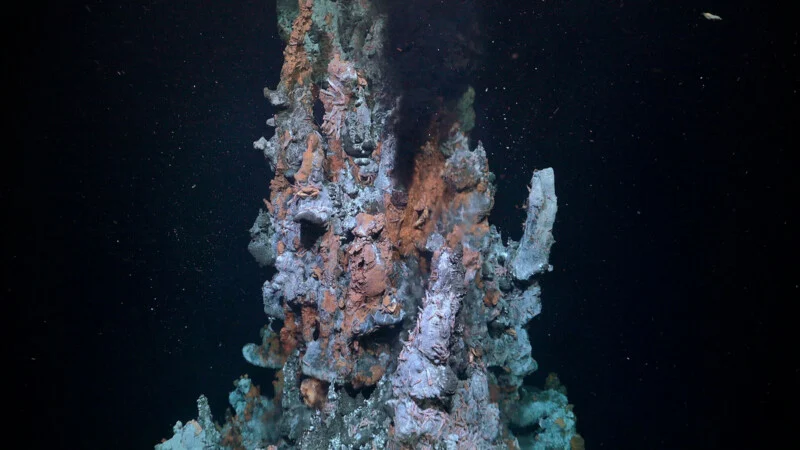 A tall underwater hydrothermal vent emits dark smoke from its top, with rugged, mineral-coated surfaces and deep-sea particles floating in the surrounding dark water.