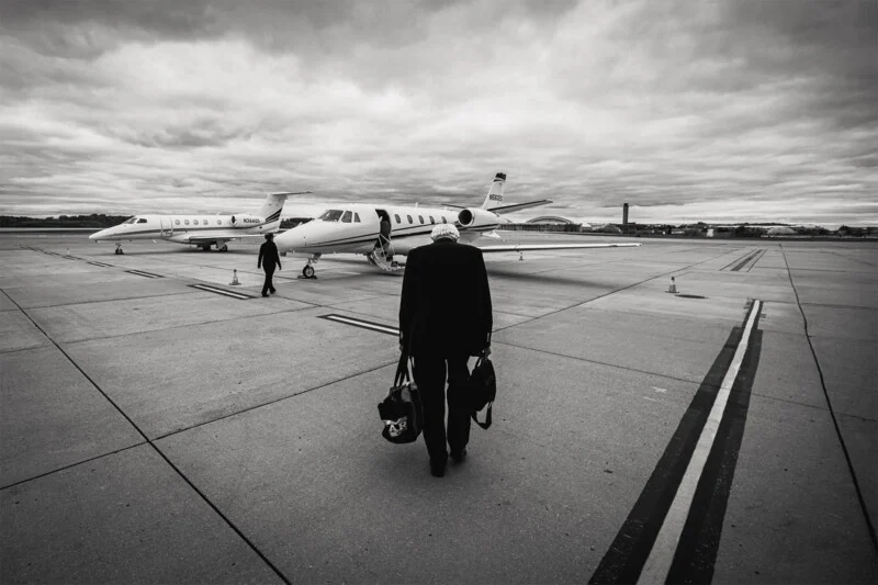 A person carrying bags walks toward a private jet parked on an airport runway, with another jet and figure in the background under a cloudy sky.