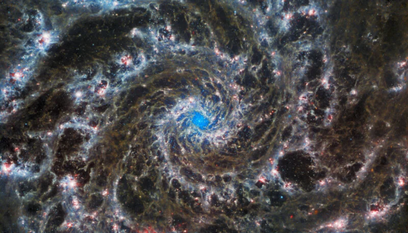 An image of the Phantom Galaxy taken by the James Webb Space Telescope. The galaxy features spiral arms displaying dust lanes, bright star clusters, and dark regions. The core glows brightly, surrounded by intricate patterns of light and dark.