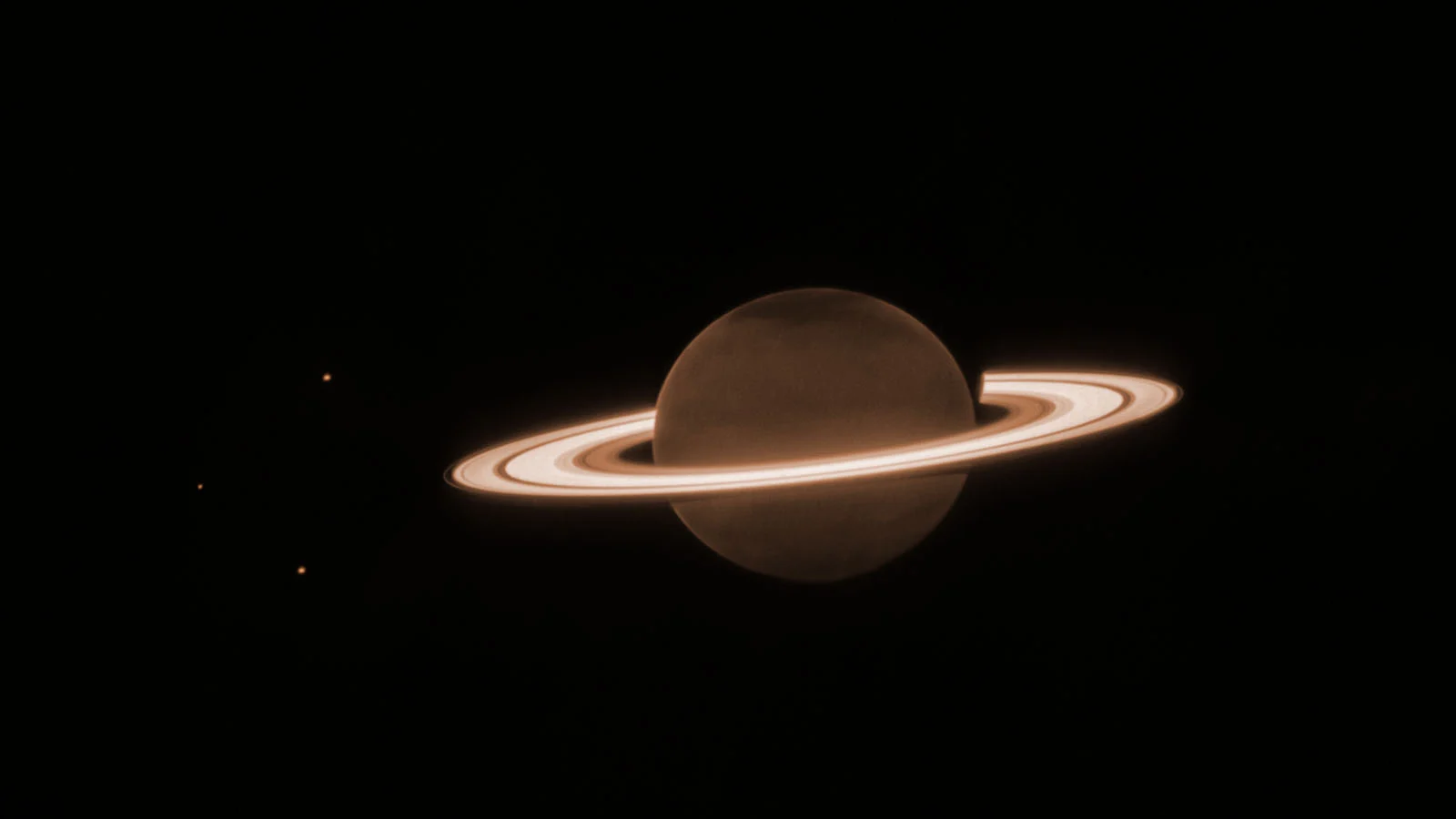 Saturn with its bright rings is seen against a dark background. Three small, faint dots, likely moons, appear to the left of the planet. The overall color tone is a muted brownish-orange.