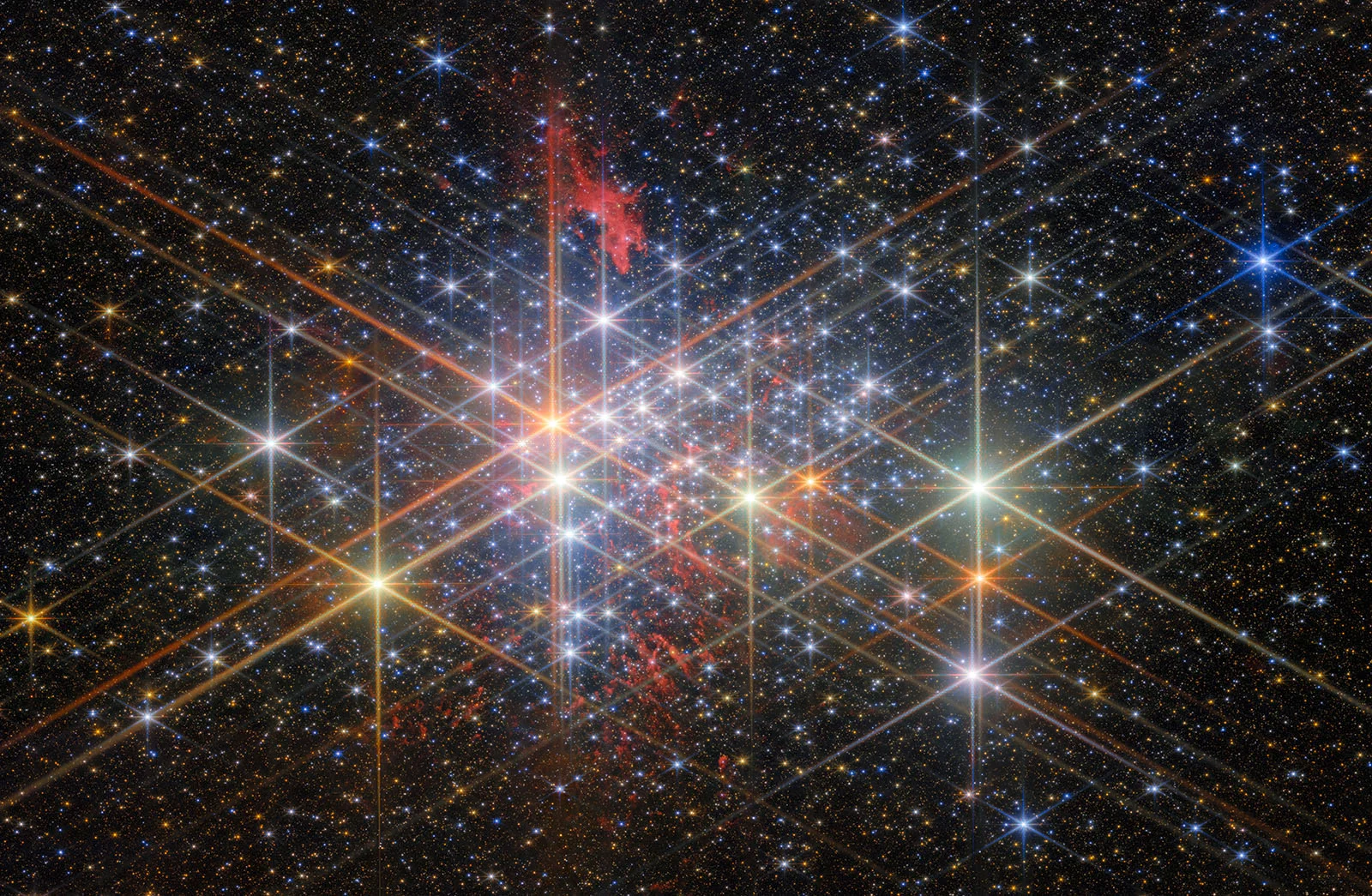 A dense star cluster sparkles with bright, colorful stars and diffraction spikes against a dark sky, with a reddish nebula visible in the upper left.