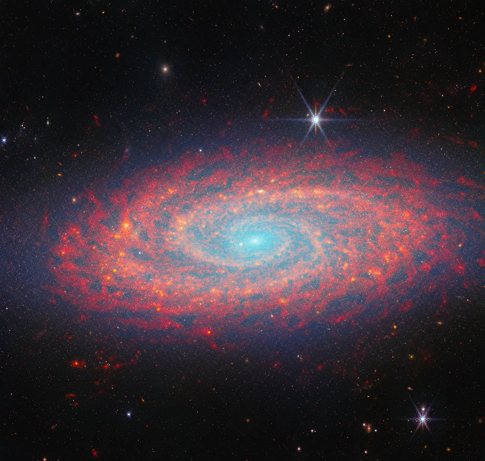 A colorful spiral galaxy glows with blue, pink, and red hues against a dark star-filled sky. Bright stars with diffraction spikes appear in the foreground.