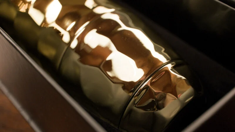 A close-up of a shiny, metallic gold cocktail shaker resting in a black box, reflecting surrounding light and colors.
