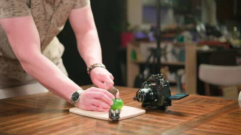 A person slices a lime on a wooden cutting board, with a digital camera and a small microphone placed nearby on the table.