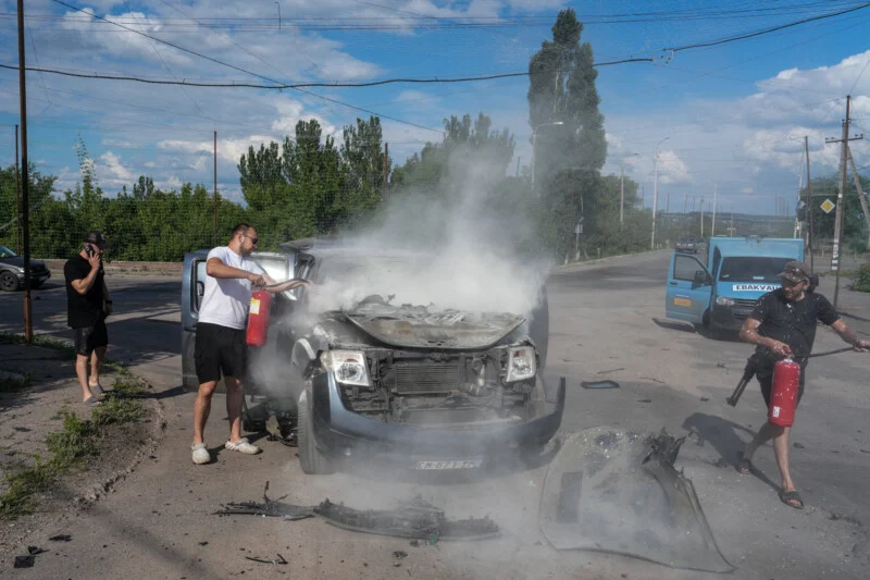 Three people use fire extinguishers to put out smoke coming from the engine of a damaged vehicle on a street, with debris scattered around and trees in the background.