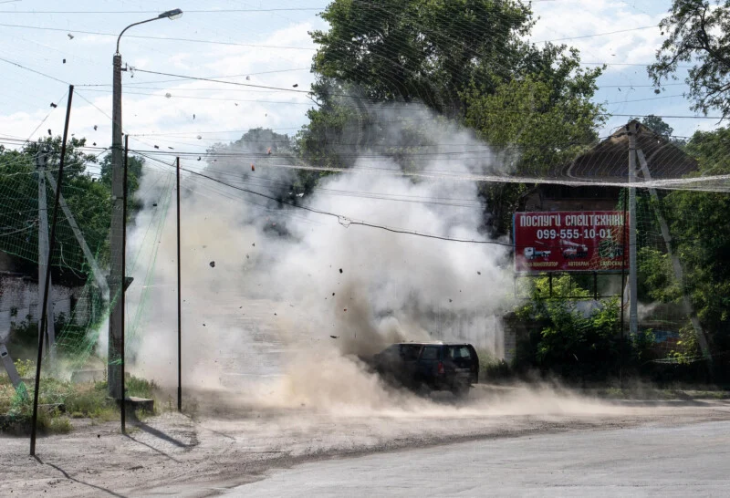 A car is engulfed in smoke and debris after an explosion on a city street, with dust and fragments in the air. Trees and a billboard are visible in the background.