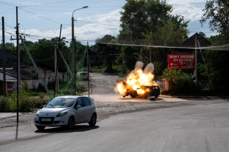 A burning car explodes on a deserted street as a white car drives by. Trees, power lines, and a red billboard are in the background on a sunny day.
