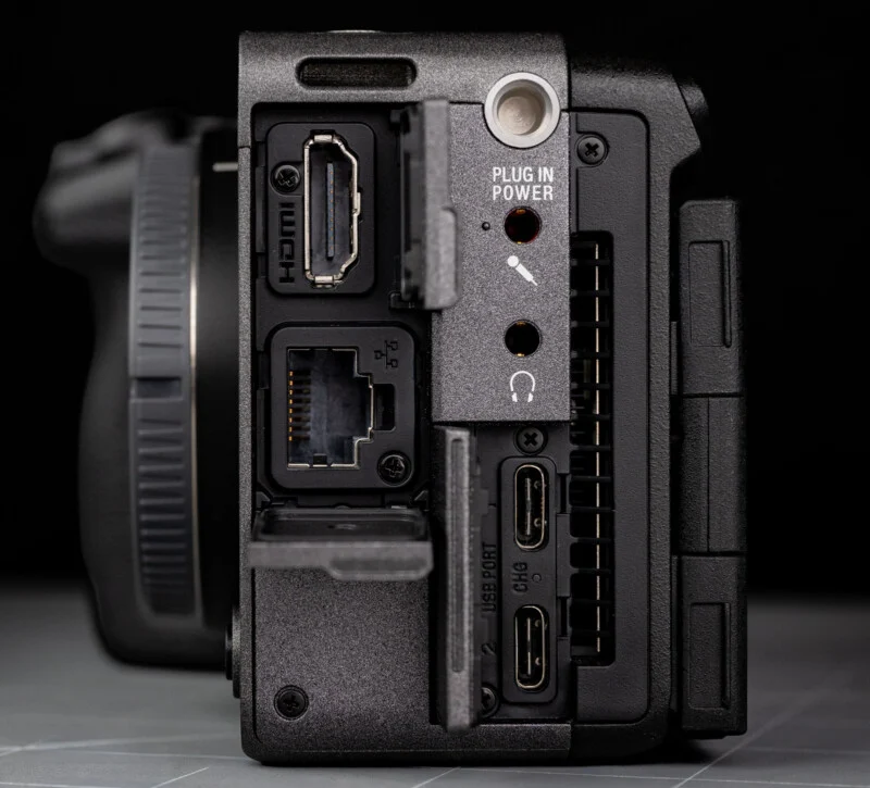 Close-up of a camera's side panel showing various ports: HDMI, LAN, USB-C, headphone and microphone jacks, and a power input. The covers for some ports are open, revealing the connectors.