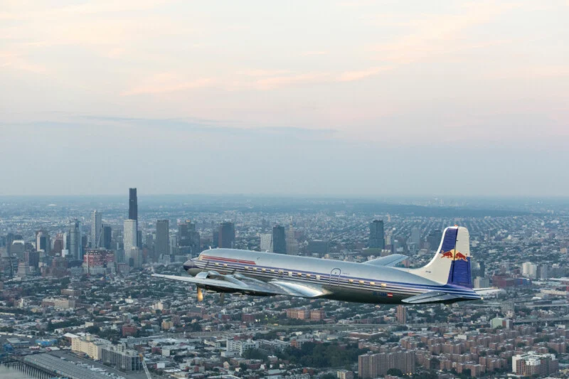A vintage silver airplane with red and blue accents flies over a sprawling cityscape with tall buildings and a cloudy sky in the background.