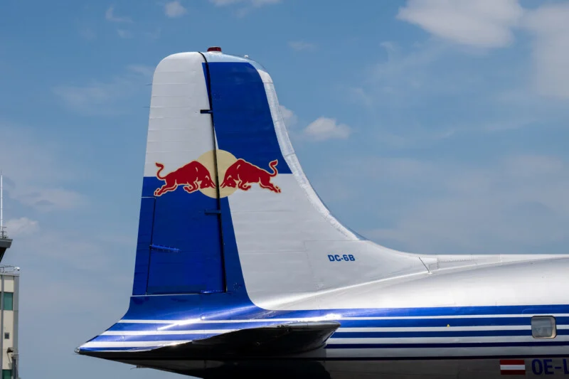 Tail section of a silver and blue aircraft with a Red Bull logo on the fin, against a partly cloudy sky. The aircraft registration "DC-6B" is visible.