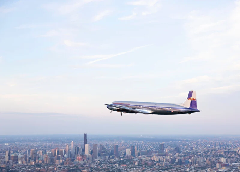 A vintage propeller airplane flies above a sprawling city skyline under a blue sky with light clouds.