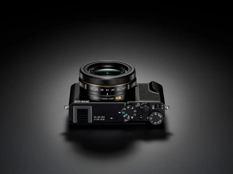 A sleek black Nikon compact camera with a lens marked “NIKKOR,” shown from above on a dark surface. The top dials, lens, and buttons are clearly visible.