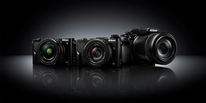 Three Nikon cameras of varying sizes and lens types are displayed side by side on a glossy black surface with a dark, gradient background.