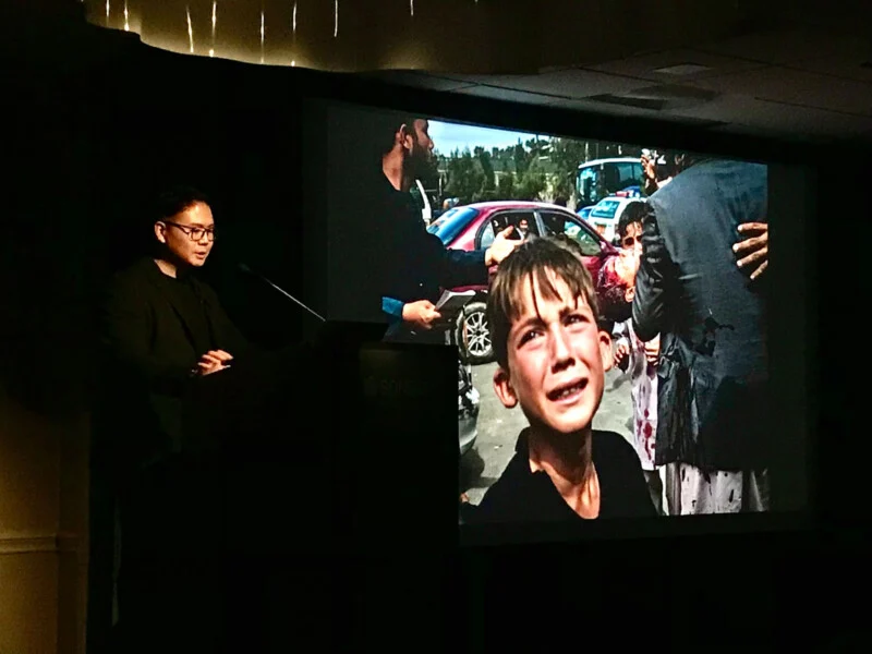A man in glasses and a dark blazer stands at a podium, speaking in front of a large screen displaying a photograph of a crying boy amidst a scene of conflict.