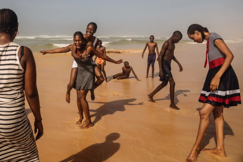 A group of people play and laugh together on a sandy beach, with some standing in the shallow water and one person giving another a piggyback ride. The ocean waves are visible in the background.