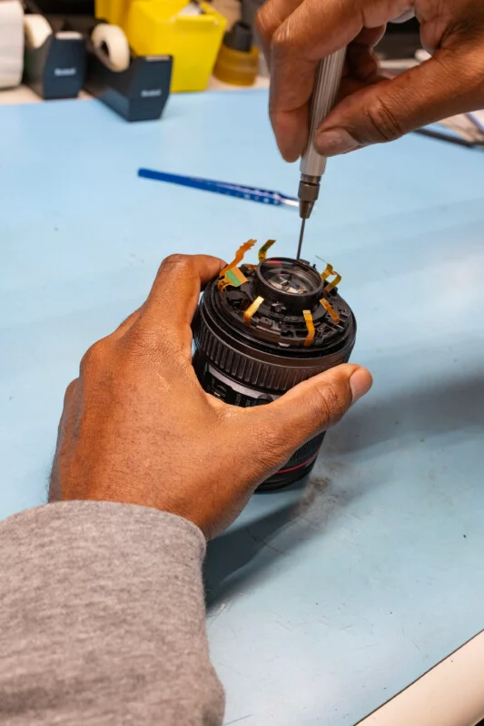 A person uses a precision screwdriver to work on the internal components of a disassembled camera lens on a blue workbench.