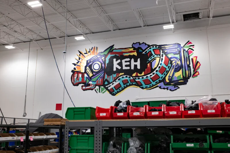 A colorful mural featuring the letters "KEH" surrounded by stylized camera and film strip imagery on a white warehouse wall above rows of storage bins.