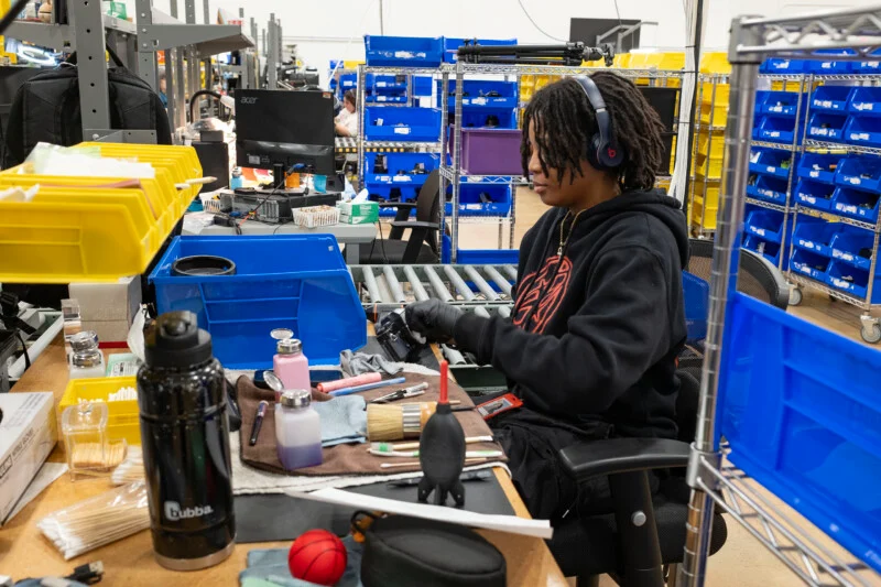 A person with dreadlocks and headphones works at a desk in a warehouse, cleaning a camera lens with tools and supplies spread out before them.