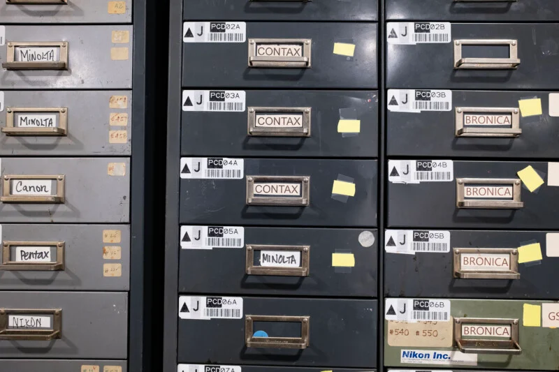 A metal filing cabinet with multiple drawers labeled with brand names including Minolta, Contax, Bronica, Canon, Pentax, and Nikon.