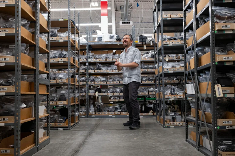 A man in a plaid shirt and dark pants stands in a warehouse aisle, laughing and clapping his hands between tall metal shelving units filled with boxed and bagged items.