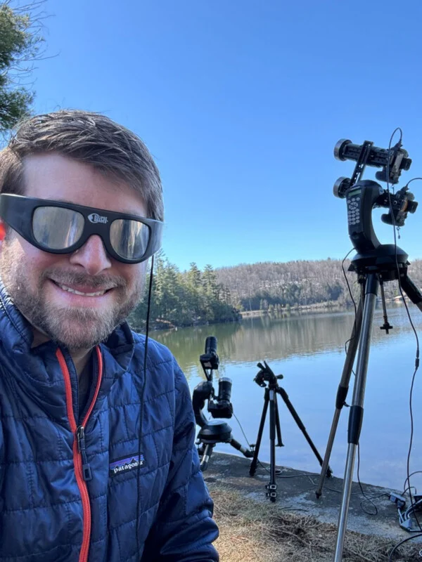 A man wearing eclipse glasses smiles for a selfie outdoors by a lake, with two camera tripods set up behind him.