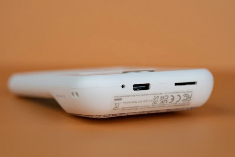 A white digital camera rests on an orange surface, showing its bottom edge with a USB-C charging port, a memory card slot, and regulatory text.
