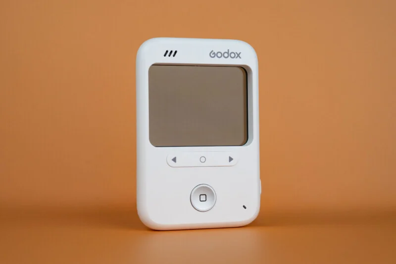 A white Godox wireless flash trigger with a blank screen, directional buttons, and a central circular button, set against a solid orange background.