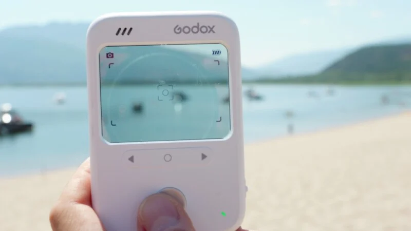 A hand holds a white Godox device with a digital screen displaying a camera viewfinder interface against a blurred beach and water background.