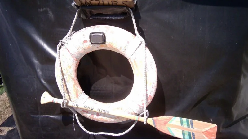 A weathered life ring hangs against a black background, with a wooden paddle featuring a painted geometric design resting horizontally across it.
