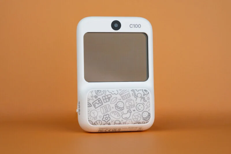 A white, rectangular Godox C100 transparent viewfinder camera with a patterned lower section sits centered against a solid orange background.