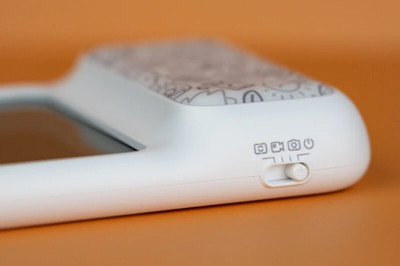 A close-up of a white device with a sliding switch, showing icons for power, camera, video, and a square frame on an orange background.