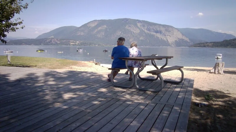 An older couple sits at a wooden picnic table on a deck overlooking a lake with mountains in the background.