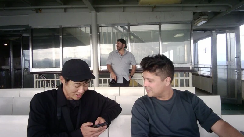Two men sit on a ferry bench while a third man stands behind them holding a camera.
