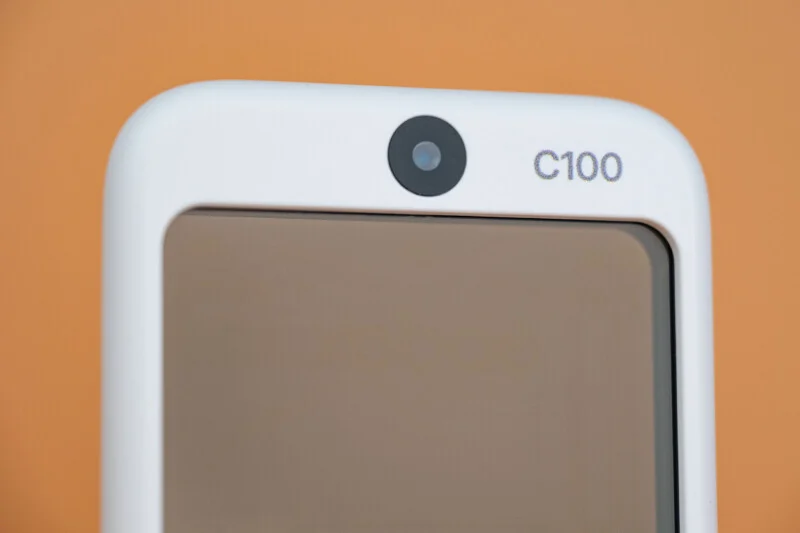 A close-up of a top portion of a white electronic device featuring a centered circular camera lens and the text "C100" printed to its right against a solid orange background.