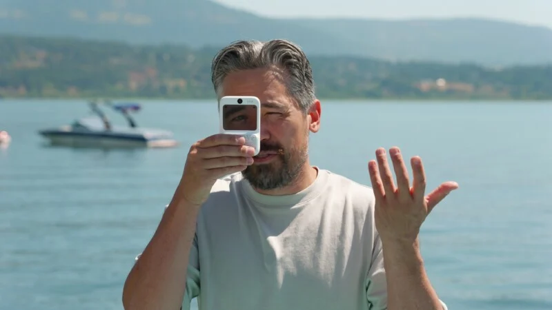 A man with graying hair and a beard holds a small white digital camera in front of one eye while gesturing with his other hand, with a lake and boat in the background.