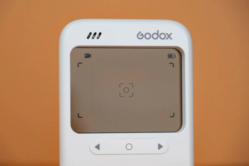 A close-up of a white Godox device with a screen displaying a video icon, a battery indicator, and a central focus frame, positioned against a solid orange background.