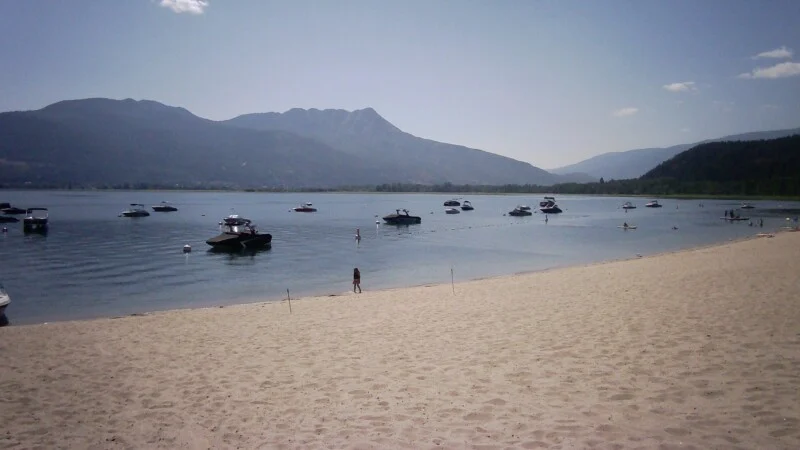 A sandy beach borders a calm lake with numerous motorboats anchored offshore under a clear blue sky and distant mountains.