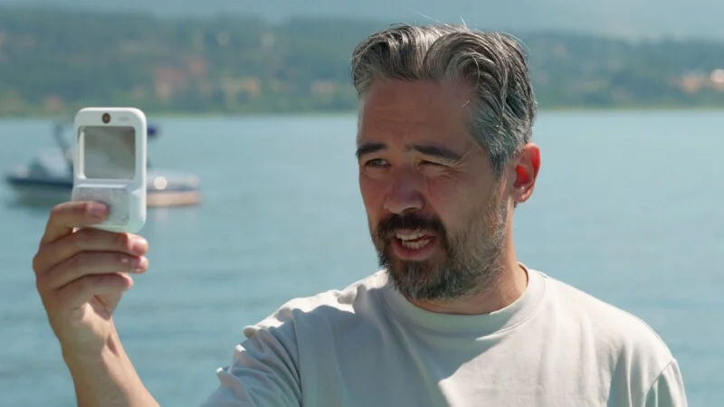A middle-aged man with graying hair and a beard squints while holding a small, white rectangular device in front of his face outdoors by a lake.